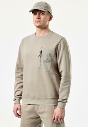 Beige genser med glidelås på brystlommen og logodetaljer. Laget av mykt stoff, med ribbestrikkede mansjetter og nederkant. Kombinert med matchende bukser.