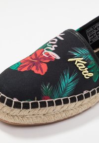Zapato negro tipo slip-on con patrones florales coloridos y "Karl" bordado en amarillo. Un ribete de cuerda de yute contorna la suela.