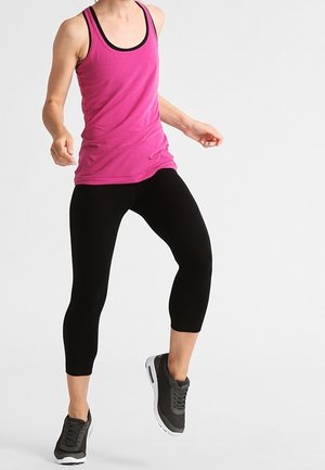 Różowy sportowy top na ramiączkach z czarnym wykończeniem, w połączeniu z czarnymi legginsami 3/4. Modelka ma na sobie szare sneakersy i przyjmuje dynamiczną pozę.