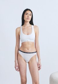 Fehér sportmelltartó és tanga szett ellentétes márkajelzésekkel az elasztikus szalagokon, sima textúrával és testhezálló kialakítással.