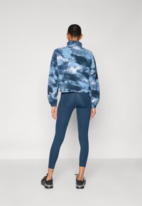 Blå tie-dye fleece pullover med høj krave, parret med mørkeblå leggings. Modellen har sorte snørebåndssko på.