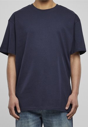 Man in een eenvoudig donkerblauw oversized t-shirt en vervaagde blauwe jeans, staande met zijn armen ontspannen langs zijn zij tegen een grijze achtergrond.