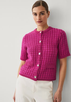 Woman wearing a textured pink short-sleeve cardigan with white buttons and white pants, standing against a plain light gray wall.