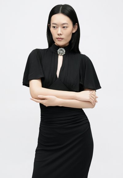 Fekete ruha rövid, bővülő ujjakkal és elején kulcslyuk alakú nyílással, melyet a nyakkivágásnál díszes ezüst bross díszít. Sima, testhezálló anyagból.