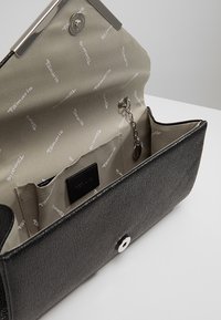 Musta teksturoidulla nahkakassilla, jossa on hopeinen napitus ja vaaleanbeige vuori, jossa on toistuva logokuviot. Mukana tulee pieni ketju.