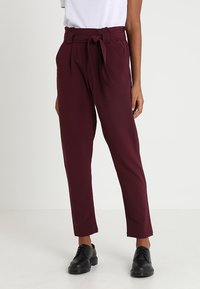 Bordeauxkleurige broek met hoge taille, strikceintuur, plooien en taps toelopende pijpen; gemaakt van een gladde stof met een matte afwerking. Zwarte schoenen worden gedragen.