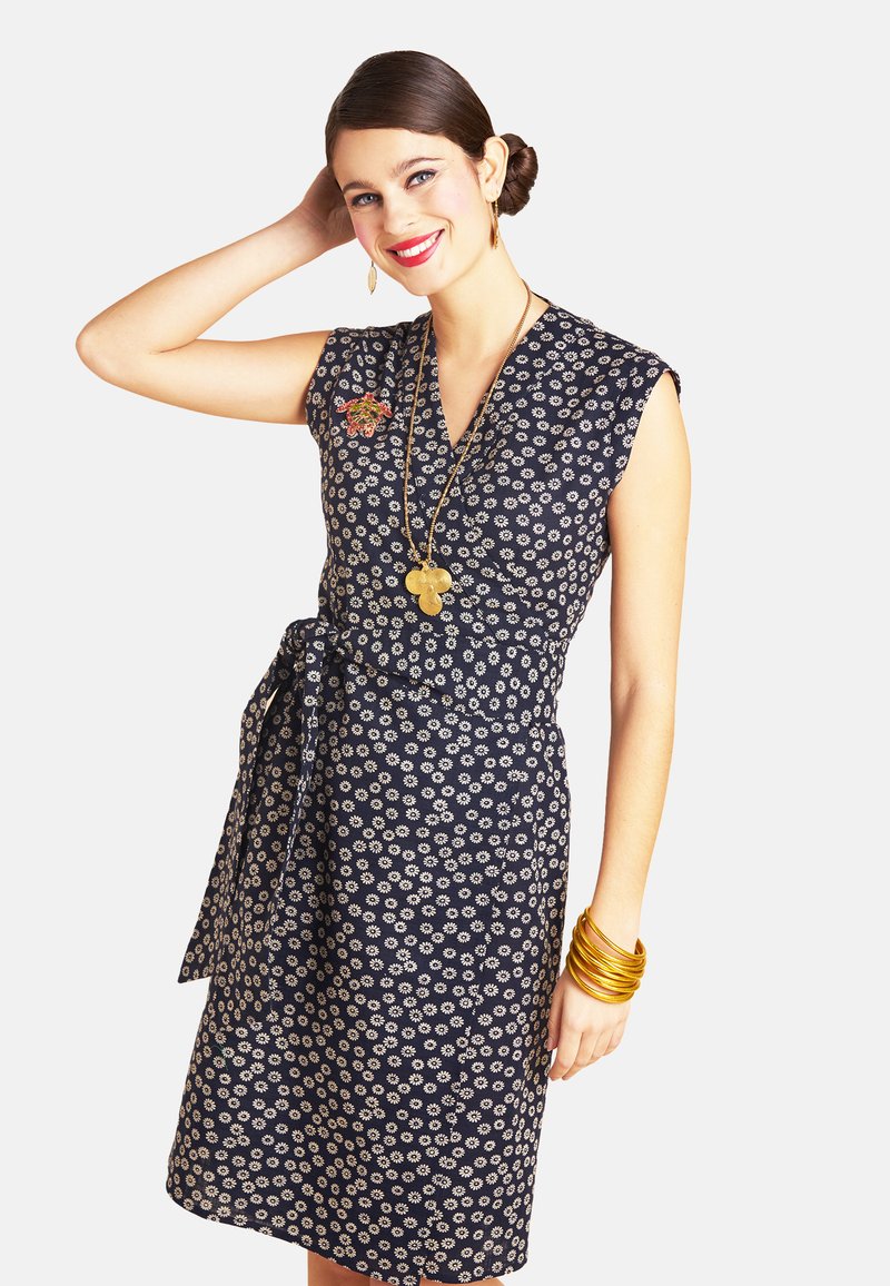 Marineblaues Kleid mit weißen Blumenmustern, kurzen Ärmeln, Wickeldesign und gebundenem Taillengurt. Accessoires umfassen goldene Armreifen und eine lange Halskette.