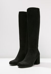 Para czarnych zamszowych kozaków do kolan z grubymi obcasami, okrągłymi noskami i zamkami błyskawicznymi na boku, na białej powierzchni.