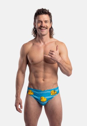 Uomo sorridente senza maglietta con mullet e baffi, che indossa un costume da bagno blu con motivi di paperelle gialle, che fa il gesto della mano shaka.