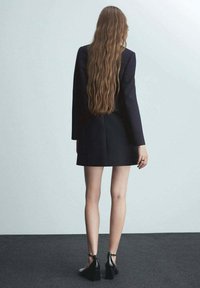 Black blazer dress with long sleeves; structured shape; mid-thigh length; paired with black heeled shoes; long wavy hair visible at the back.