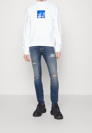 Una persona está de pie vistiendo un sudadera blanca con un logo azul, jeans azules desgastados y zapatillas negras en un entorno minimalista.
