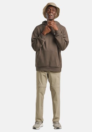 Homme souriant en sweat à capuche marron ajustant la lanière de son chapeau, portant un pantalon beige, des baskets et un bob marron clair, debout devant un fond blanc.