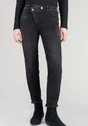 Jean en denim noir avec une coupe ajustée, présentant des détails déchirés, deux poches avant et une fermeture à bouton. Ourlets roulés au-dessus de la cheville.