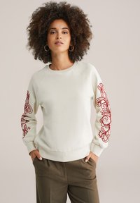 Felpa color crema con maniche lunghe, caratterizzata da intricate ricami floreali rossi su entrambe le maniche; vestibilità morbida e polsini a coste.