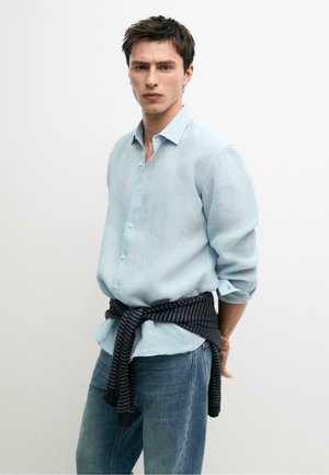 Jeune homme portant une chemise bleu clair boutonnée, un jean bleu, avec un pull rayé noir et gris noué autour de sa taille.