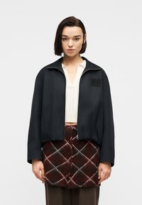 Γυναίκα με κοντά σκούρα μαλλιά φοράει μαύρο μπουφάν με φερμουάρ πάνω από λευκή πλεκτή μπλούζα και καφέ καρό φούστα με κουμπιά, στέκεται σε απλό φόντο.