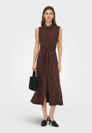 Donna in piedi che indossa un abito midi marrone senza maniche con cintura, mocassini neri con tacco, che tiene una borsa nera contro uno sfondo semplice.