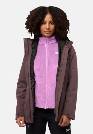Femme aux cheveux bouclés portant une veste en polaire violette partiellement zippée sous un imperméable marron, se tenant devant un fond uni.