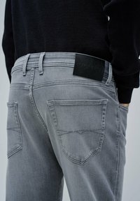 Jeans de mezclilla gris con un ajuste elegante, que presentan una etiqueta de cuero negro en la cinturilla y bolsillos traseros estándar con detalles de costura sutiles.