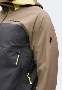 Veste bicolore marron et grise avec un logo noir sur la manche et le texte Peak Performance sur la poitrine, avec des poches zippées à tirettes jaunes.