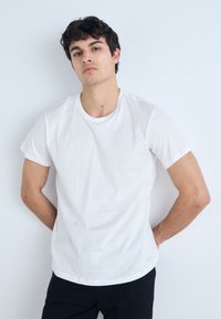 White cotton t-shirt with short sleeves, crew neckline, and a straight hem. Worn with black pants, featuring a subtle logo detail at the bottom.
