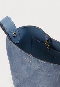 Błękitna zamszowa torba typu 'bucket' marki Coach z skórzanym paskiem, złotymi okuciami oraz widoczną wewnętrzną kieszenią zamykaną na zatrzask.