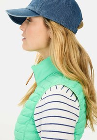Helle grüne Pufferweste mit hohem Kragen, getragen über einem weiß-blau gestreiften Langarmoberteil. Dunkelblaue Denim-Mütze. Langes, welliges blondes Haar.