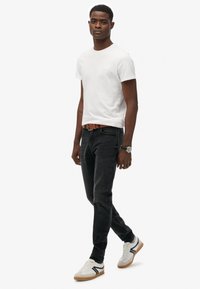 Biała krótka koszulka z krótkim rękawem, ciemne dopasowane dżinsy, brązowy pasek i białe trampki z czarnymi akcentami. Model stoi na neutralnym tle.