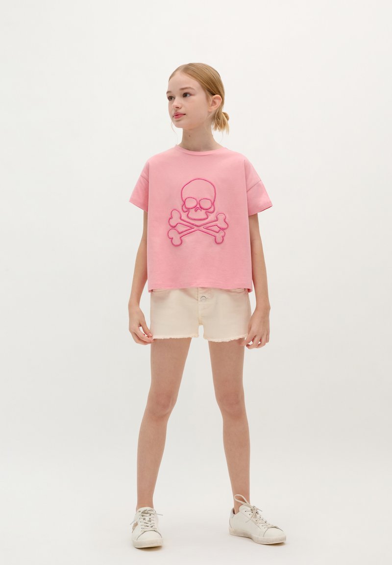 Rosa Baumwoll-T-Shirt mit einem großen gestickten Totenkopf- und Knochen-Design, kombiniert mit beigen Denim-Shorts und weißen Sneakers.