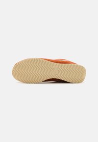 Semelle en caoutchouc beige avec motif à chevrons, logo Nike embossé au centre, fixée à une tige en suède orange de sneaker, photographiée vue de la semelle.