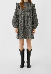 Nainen, joka on pukeutunut mustaan kukkakuosisessa mekkoon, jossa on puffihiha ja mustat polvipitkät cowboy-saappaat, seisoo yksinkertaista taustaa vasten.