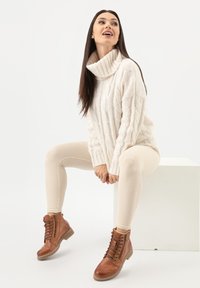 Maglione a trecce di crema spesso, leggings beige a coste e stivaletti alla caviglia marroni con suole testurizzate, seduto su uno sfondo bianco.