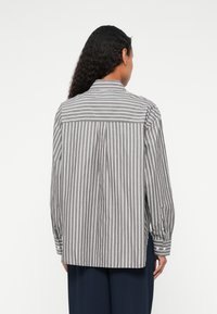 Camicia a righe grigie e bianche con yoke posteriore, maniche lunghe, polsini con bottoni e orlo dritto. Il tessuto sembra intrecciato e texturizzato.