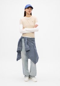 Beige tettskjorte med fasong laget over en hvit langermet topp, kombinert med lyseblå denimbukser og en blå rutete skjorte knyttet rundt livet. Blå caps.