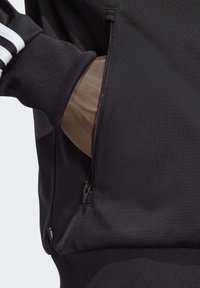 Svart sportjacka med texturerad tyg, dragkedjefickor och randiga detaljer på ärmarna.