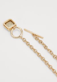 Accessoire en chaîne dorée avec un pendentif hexagonal arborant un logo, un anneau circulaire et un accent matériel plat pour l'attache.