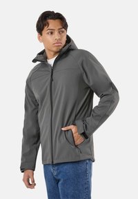 Jeune homme portant une veste softshell grise à capuche avec une fermeture éclair et les mains dans les poches, associée à un jean bleu, regardant vers sa gauche.