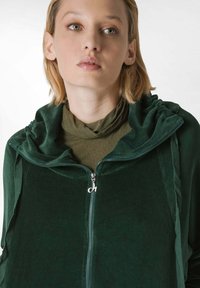 Felpa zip-up verde scuro con una consistenza morbida, tessuto arricciato sul cappuccio e una cerniera argentata. Indossata sopra un top aderente oliva.