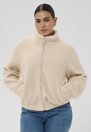 Kvinde med mørkt hår iført en cremefarvet fleecejakke med høj krave og blå jeans, hænderne i lommerne, foran en ensartet grå baggrund.