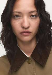 Veste marron en velours côtelé avec un tissu lisse de couleur marron clair. Le modèle a des cheveux ondulés et foncés, ainsi que des taches de rousseur naturelles, avec une expression neutre.