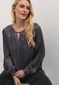 Siva satenska bluza s okruglim izrezom, detaljem u obliku ključa i dugim nabranim rukavima. Uparena s tamno sivim trapericama. Mekana tekstura i elegantan pad.