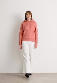Una donna sta camminando, indossando un hoodie corallo rosa, pantaloni bianchi a gamba larga e scarpe nere su uno sfondo bianco semplice.