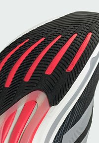 Suela de goma negra con acentos en rojo y blanco, con surcos y canales texturizados para mayor tracción. Entresuela suave gris con parte superior de malla.