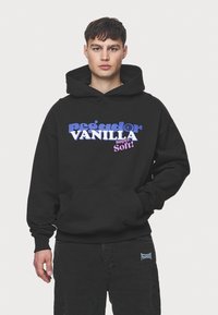 Črn hoodie s sprednjim žepom, ki vsebuje drzne napis v vijolični in roza barvi "pega dor VANILLA super Soft!" na gladki teksturi blaga.