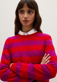 Rot-lila gestreifter Pullover mit Rundhalsausschnitt und langen Ärmeln, aus weichem Strickmaterial. Darunter ist ein weißes Hemd mit Kragen sichtbar.