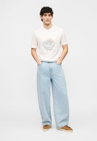 Νεαρός με σγουρά σκούρα μαλλιά που φοράει λευκό γραφικό T-shirt, ανοιχτόχρωμο μπλε τζιν παντελόνι φαρδιά γραμμή και καφέ αθλητικά παπούτσια, στέκεται με τα χέρια στις τσέπες.