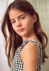 Vestito senza maniche a quadretti bianchi e neri con spalline annodate; tessuto morbido con una texture liscia; capelli ondulati e di lunghezza fino alle spalle.