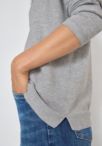 Maglione grigio lavorato a maglia con una texture a costine, caratterizzato da un collo rotondo e spacchi laterali, abbinato a jeans in denim blu.