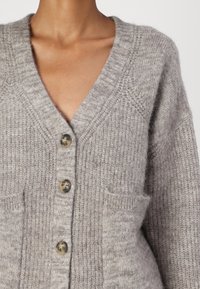 Pelēks adīts kardigans ar V-veida kaklu, ar trim lielām pogām, teksturētu audumu un divām priekšējām kabatām.