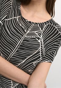 Frau trägt ein schwarz-weißes Kurzarmoberteil mit Blattmuster, die Arme verschränkt, im Bild sind Ausschnitt sowie teilweise Gesicht und Arm zu sehen.